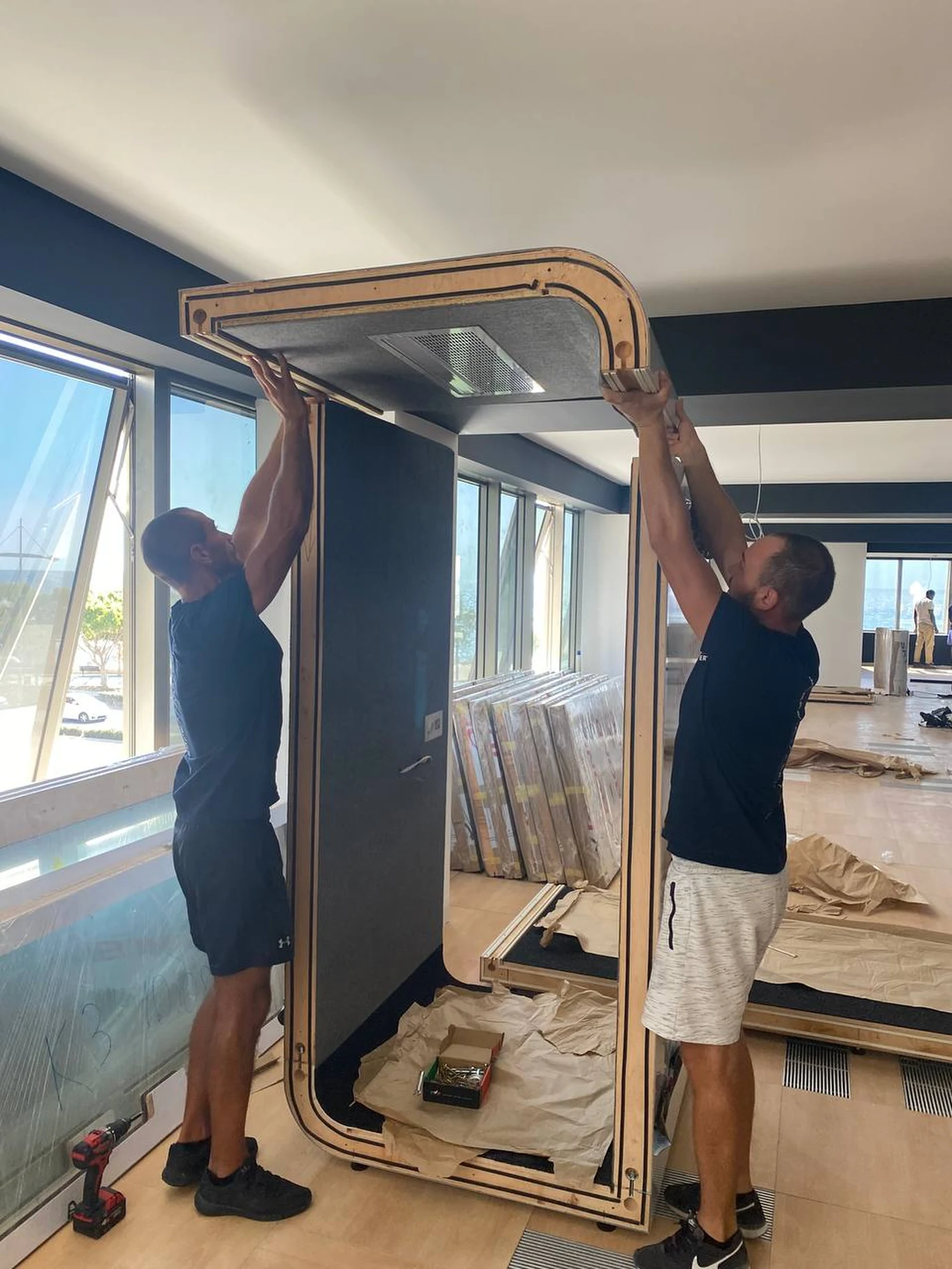Two Silentbox installers lifting and fitting the ceiling panel of a booth with a sea view